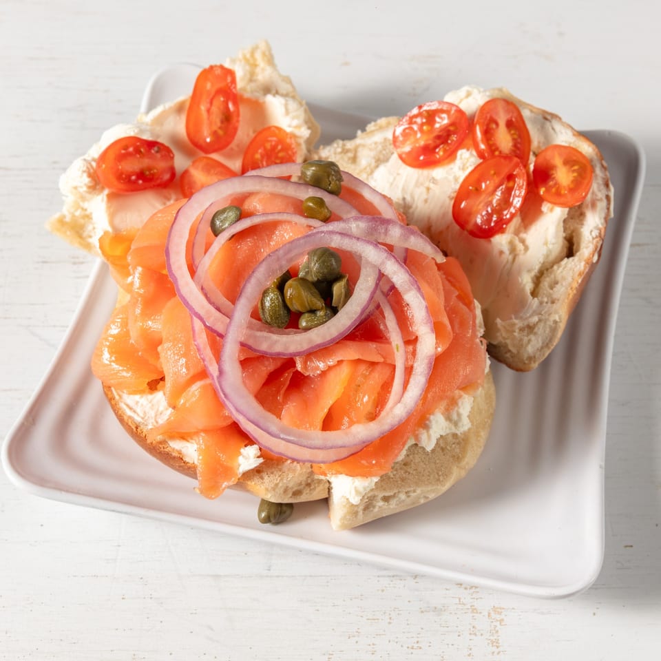 Bagel with cream cheese, smoked salmon, red onion, capers, and cherry tomatoes on a white plate.