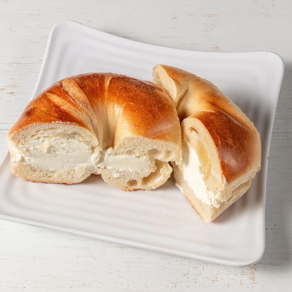 A golden-brown bagel, sliced and generously filled with cream cheese, rests on a white plate.