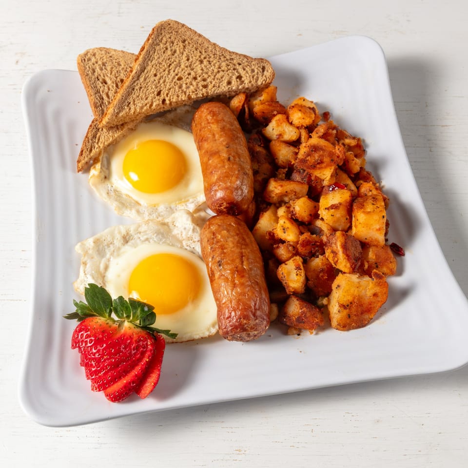 Breakfast platter with two fried eggs, two sausages, home fries, wheat toast, and a sliced strawberry.